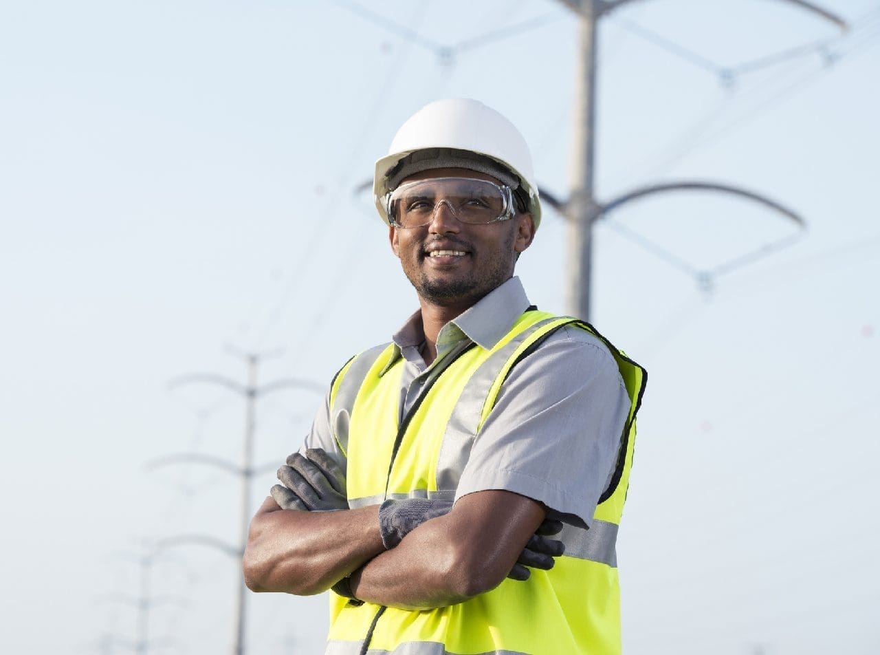 A construction worker in a reflective vest and hard hat stands confidently with arms crossed near power lines.