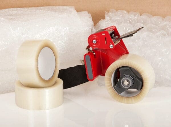 A red tape dispenser with transparent packing tape rolls and bubble wrap in the background.